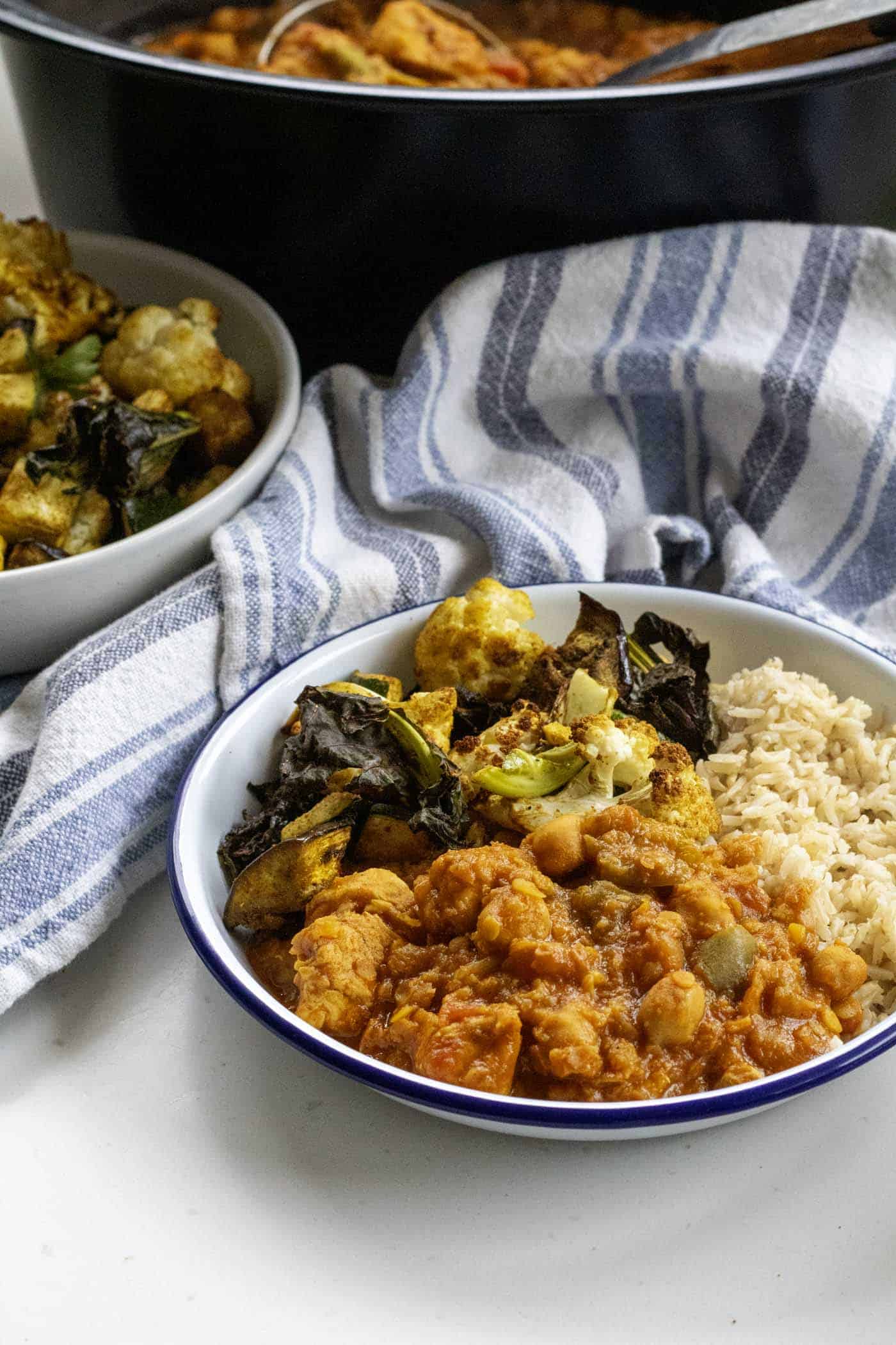 A white bowl filled with rice, vegetables, and a healthy chicken curry dish cooked in the slow cooker is placed on a white surface next to a striped cloth and another bowl containing more vegetables. the slow cooker chicken curry is in the background