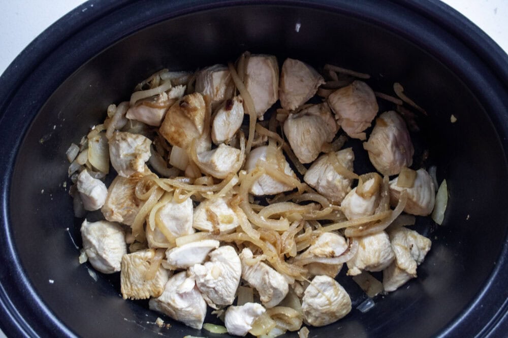 A black slow cooker bowl filled with the browned cubed chicken and onions