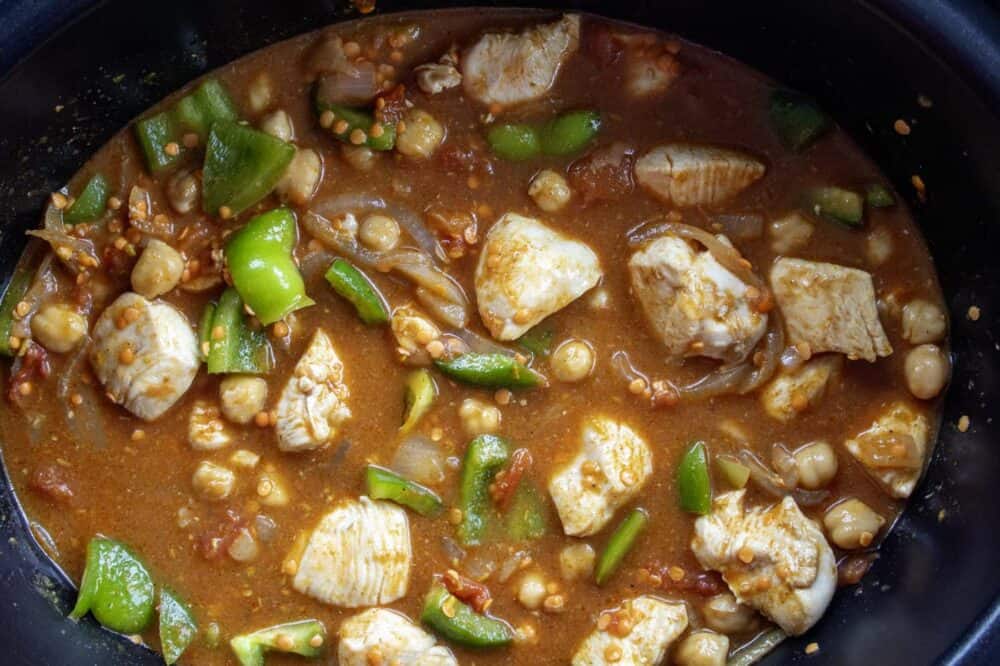 A black slow cooker bowl filled with the uncooked tomato cuury based sauce, chickpeas, lentils, cubed chicken and onions with green pepper chunks.