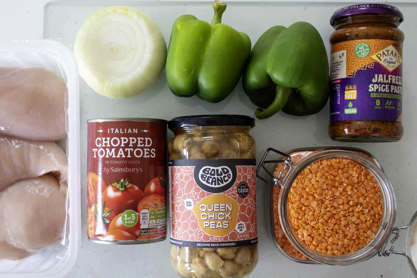 Ingredients for healthy chicken curry on a chopping board including chicken breasts, white onion, chopped tinned tomatoes, jar of chickpeas, red lentils, curry paste and 2 green peppers