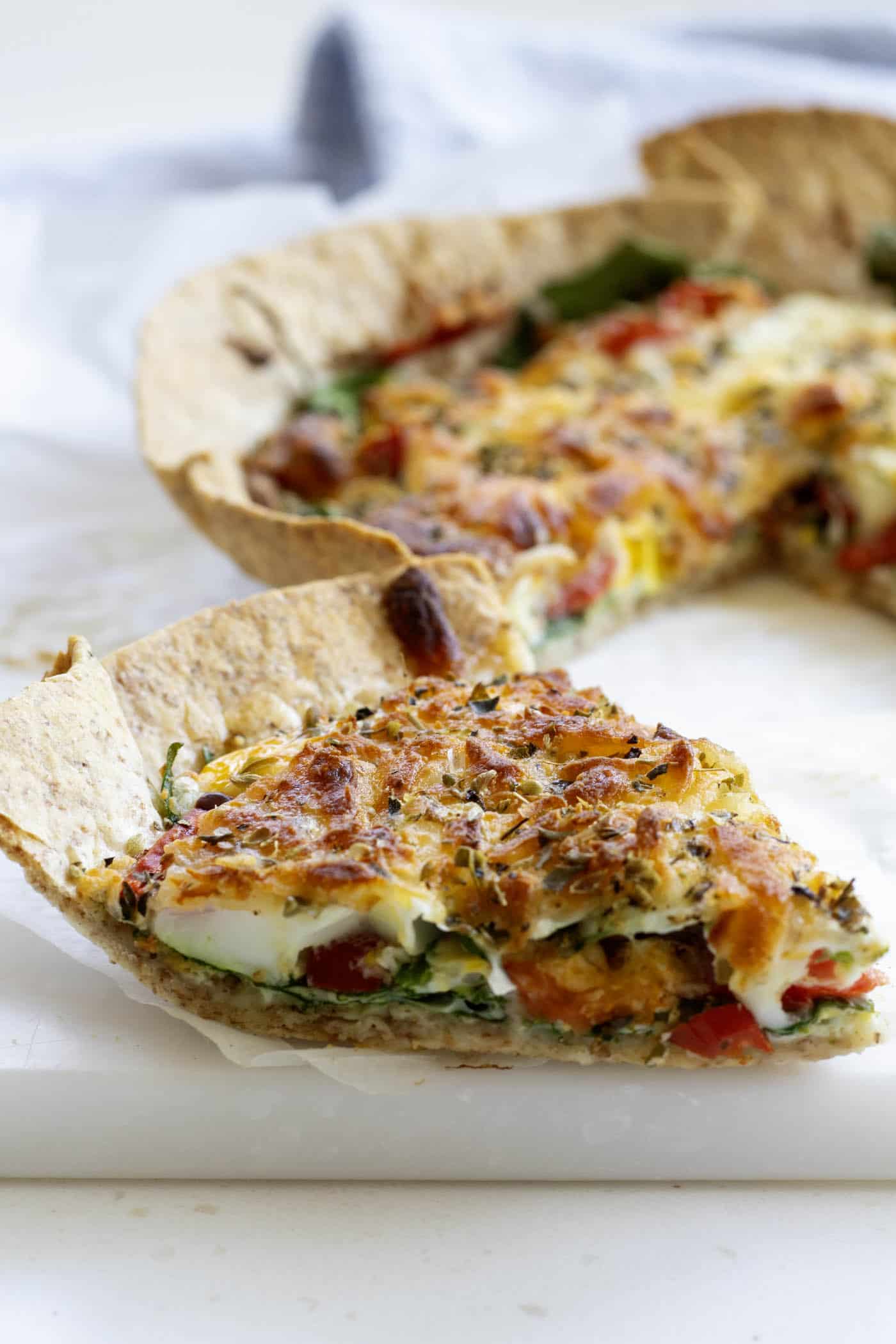 A closeup of a slice of healthy tortilla pizza on a white board and greasproof paper with a blue cloth and iced coffee in the background
