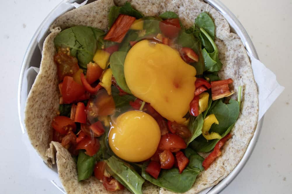 2 raw eggs cracked in a round tin stuffed with a tortilla with a layer of spinach, chopped tomatoes and peppers