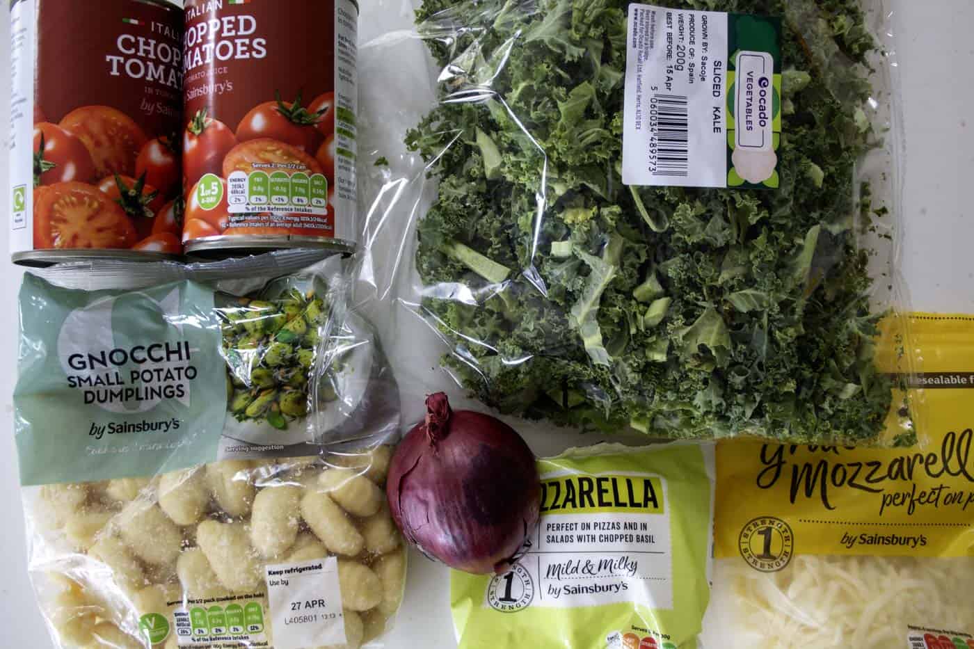 ingredients for vegetarian gnocchi bake including tinned tomatoes, a bag of kale, a bag of gnocchi, a red onion, grated and a ball of mozerella