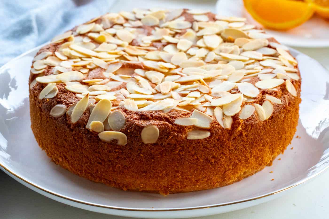 Orange almond cake on a large white cake plate with cut orange and a blue cloth in the background
