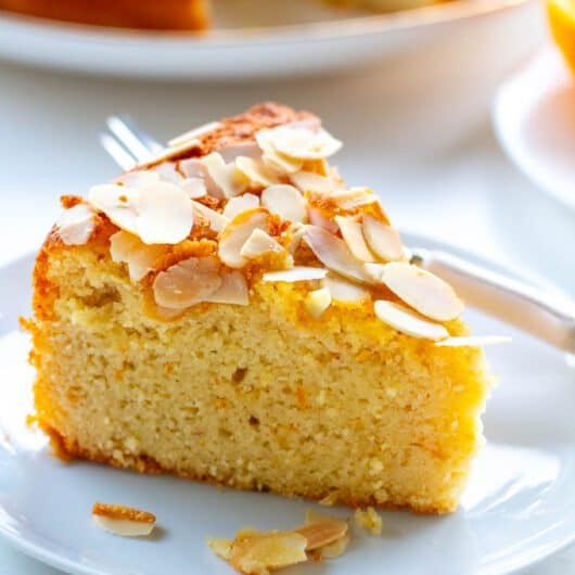 closeup of a slice of orange almond flour cake on a plate with a small fork and the cake an cut orange in the background