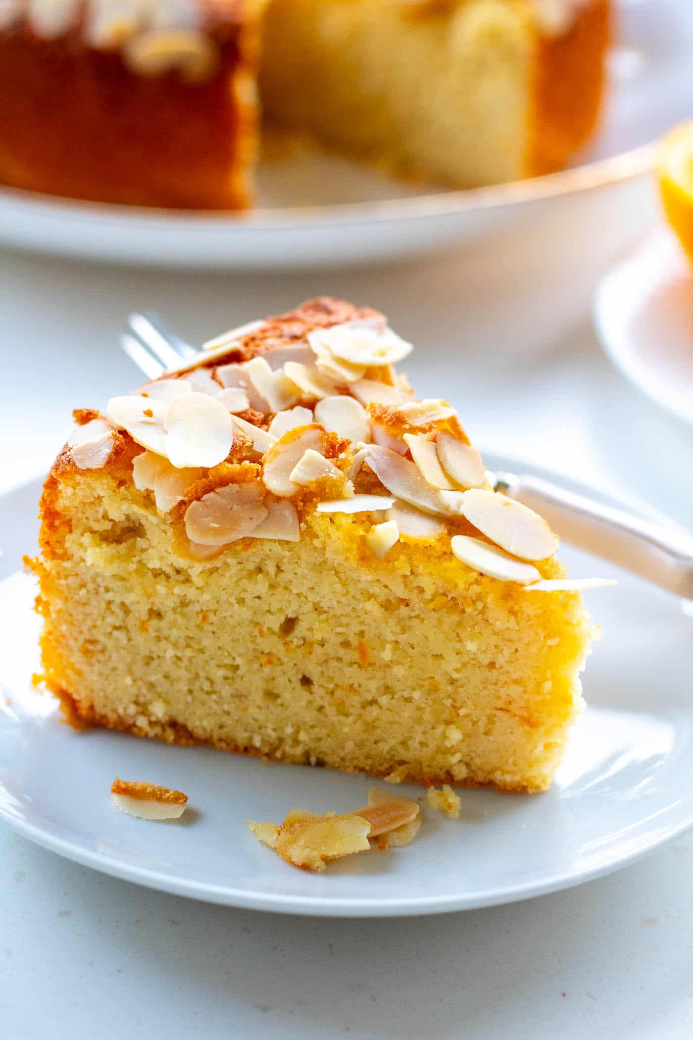 closeup of a slice of orange almond flour cake on a plate with a small fork and the cake an cut orange in the background