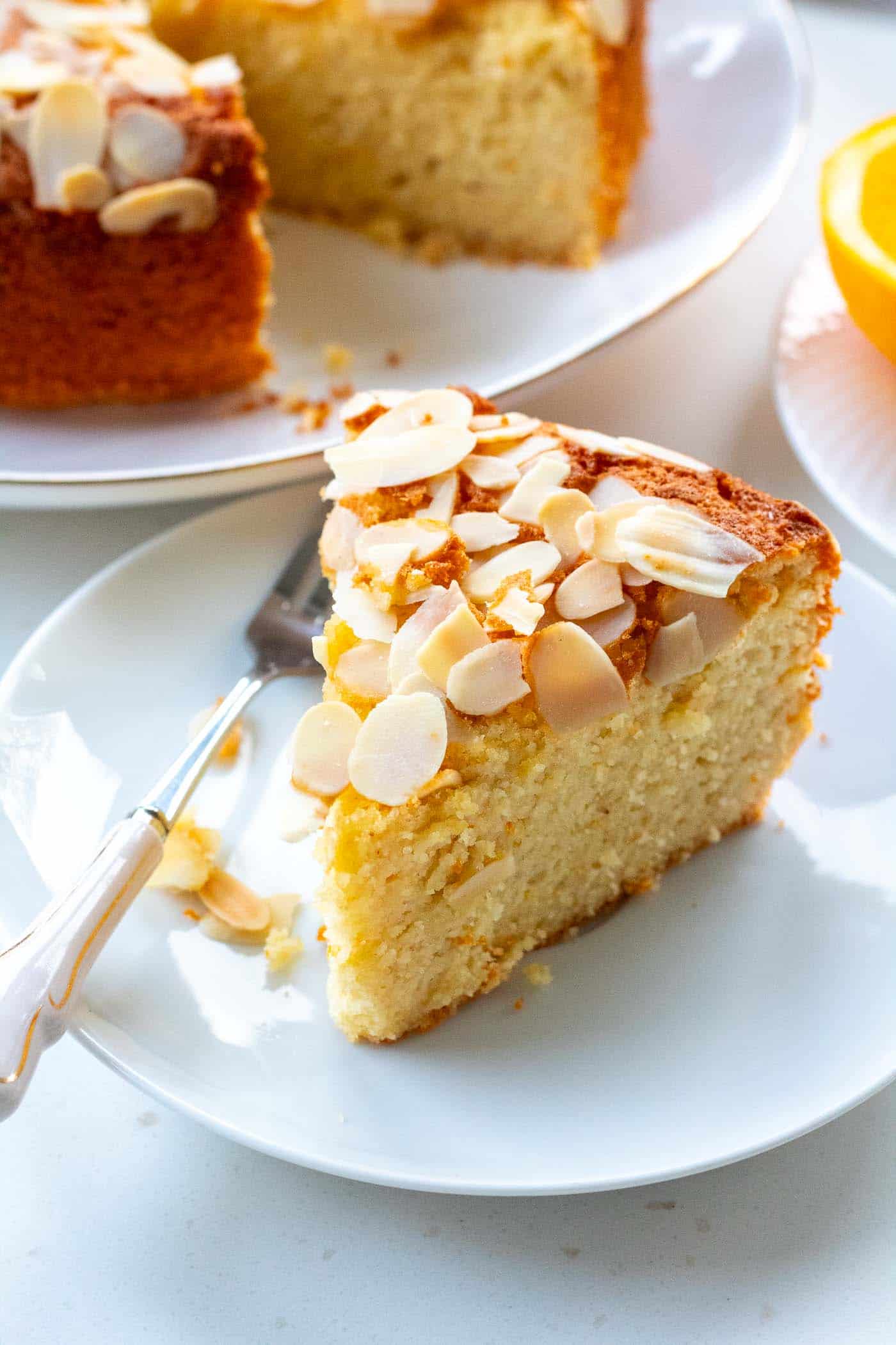 a slice of orange almond flour cake on a plate with a small fork and the cake an cut orange in the background