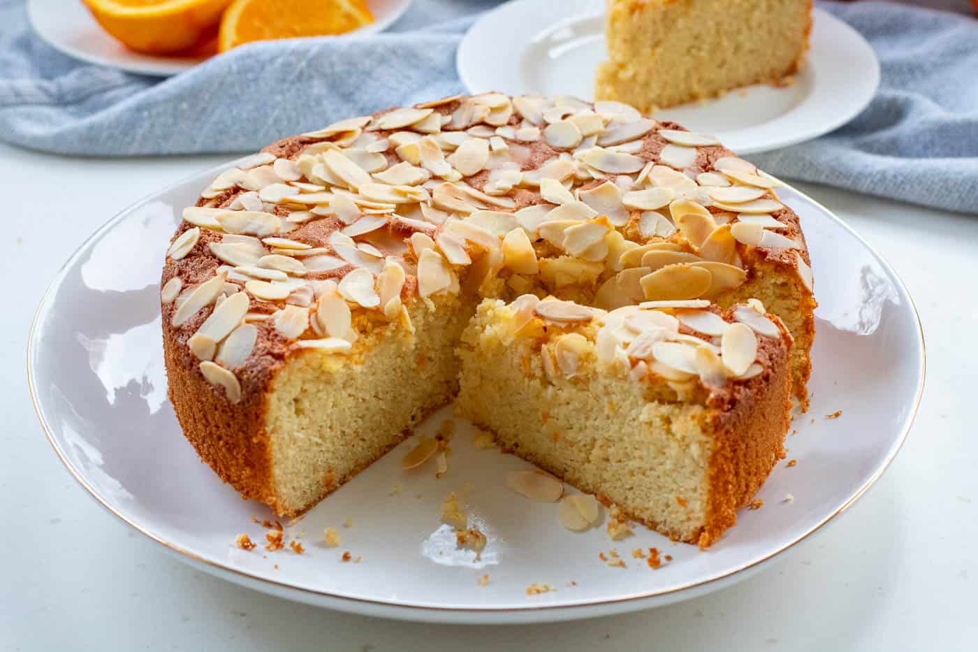 flourless orange almond cake sliced on a plate with aslice of cake and cut orange in the background on a blue cloth