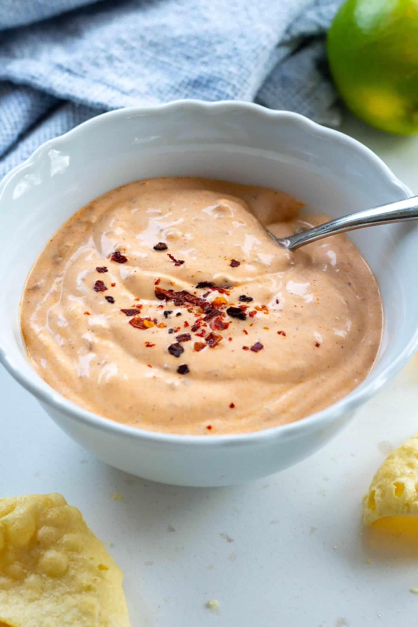 creamy chipotle sauce in a white bowl with a spoon and aleppo chillis sprinkled on top with limes, lentil crisps and a blue cloth in the background
