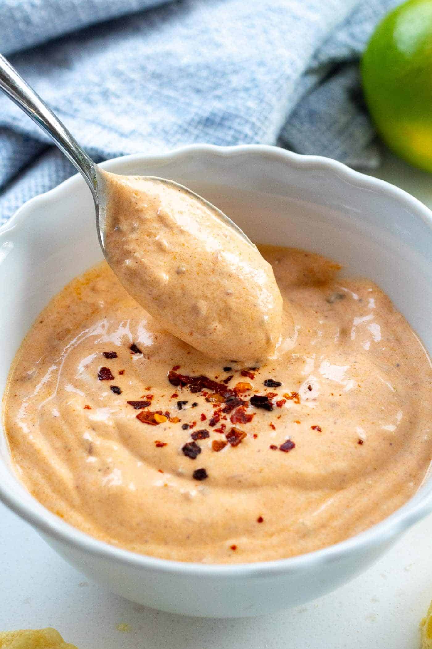 creamy greek yogurt chipotle sauce in a white bowl with a spoon lifting out and aleppo chillis sprinkled on top with limes and a blue cloth in the background