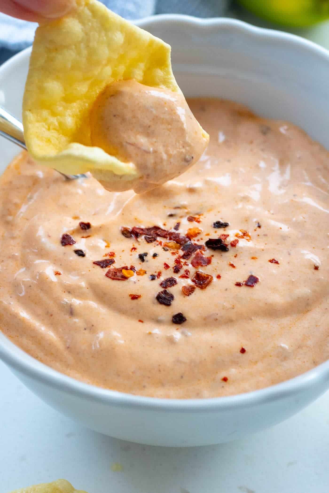 a hand dipping a tortilla chip into a bowl of chipotle dipping sauce with aleppo chillis sprinkled on top