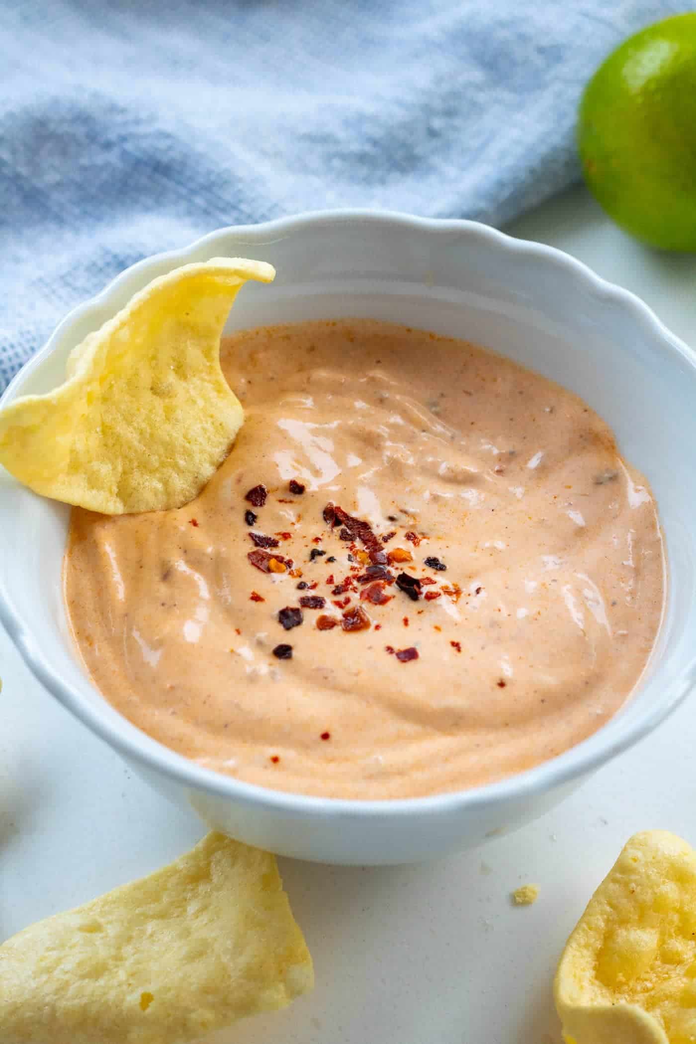 creamy chipotle sauce in a white bowl with a tritlla crisp and aleppo chillis sprinkled on top with limes, lentil crisps and a blue cloth in the background