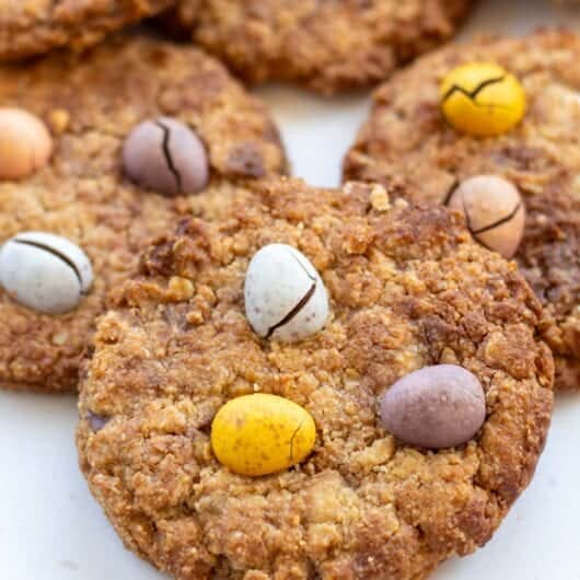 Golden-brown mini egg oatmeal cookies with a crisp edge and soft centre, topped with pastel-coloured mini eggs, set on a white surface with scattered mini eggs.