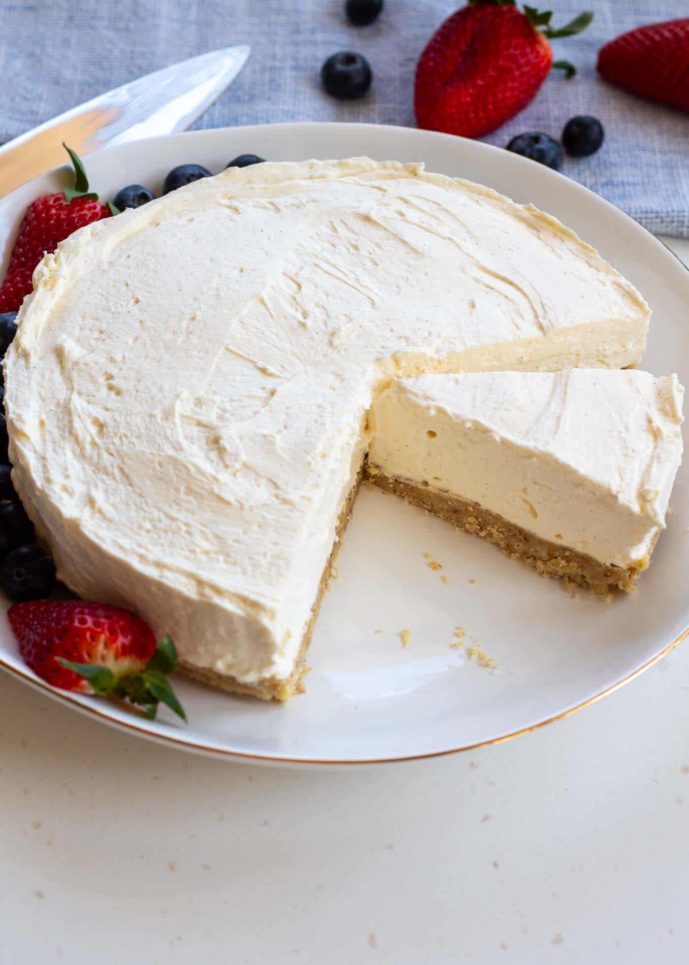 A slice taken from a sugar-free vanilla mascarpone cheesecake, highlighting its smooth, low-carb, no-bake composition. on a white plate with some strawberries and blueberries around the plate and in the background with a cake slice and blue cloth.