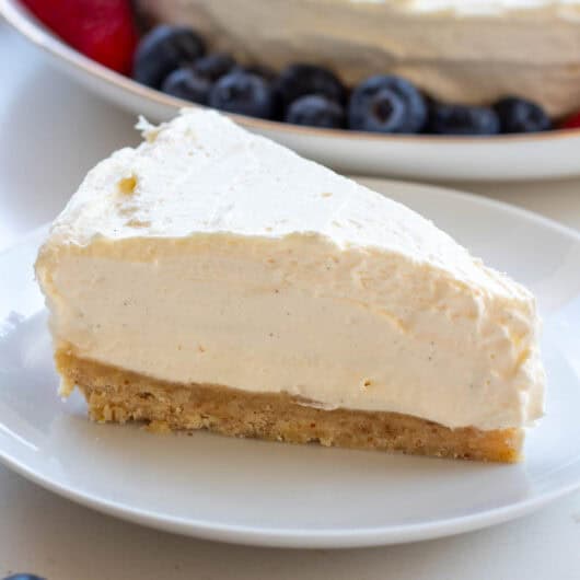 A slice of sugar-free vanilla mascarpone cheesecake elegantly presented on a simple white plate with the rest of the cheesecake and berries in the background