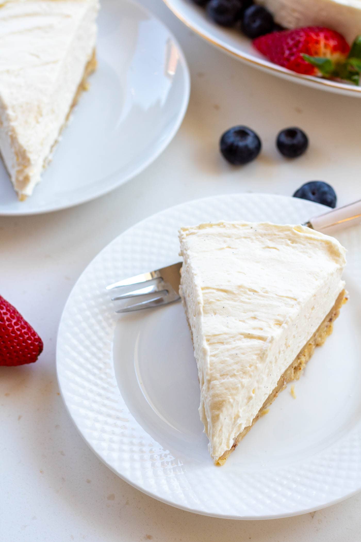 A slice of sugar-free vanilla cheesecake on a white plate with a cake fork and fresh berries and more cheesecake slices in the background