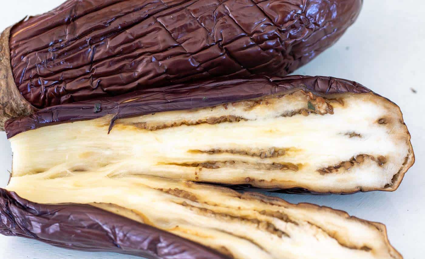 2 roasted aubergines on a white background with one sliced in half lenghtways