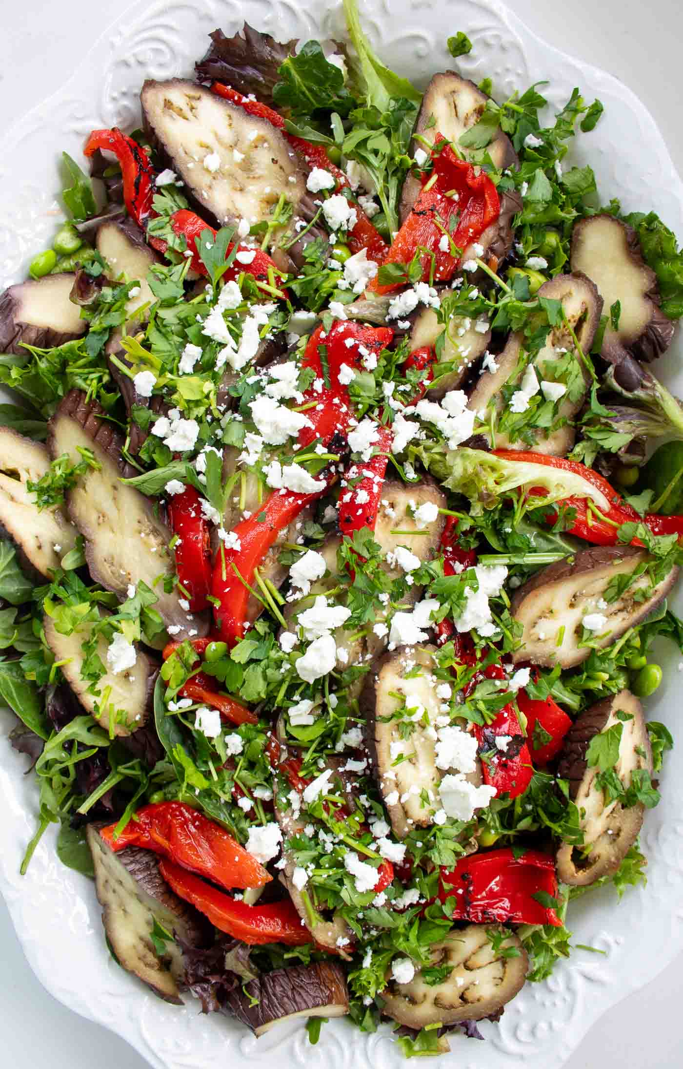 A vibrant roasted aubergine and pepper salad featuring sliced aubergines, red peppers, green beans, and crumbled feta cheese with chopped parsley on a bed of salad leaves ona decorative white platter