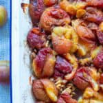 Close-up of spiced roast plums in a white baking dish, with fresh plums to teh side. An easy low-sugar dessert recipe.