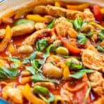 Close-up of Spanish chicken casserole in a blue cast-iron pan, with crusty seeded bread in the background, showing tender chicken, creamy butter beans, sweet peppers, and green olives in a rich smoky tomato sauce