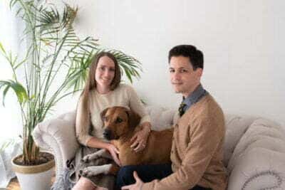 picture of Adam, his wife, and dog. They are sitting together, snuggled up on a couch.