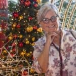 Χαμογελαστή πελάτισσα σε εορταστικό περιβάλλον κάνει τα χριστουγεννιάτικα ψώνια της, μπροστά από στολισμένο δέντρο και φωτεινές διακοσμήσεις.