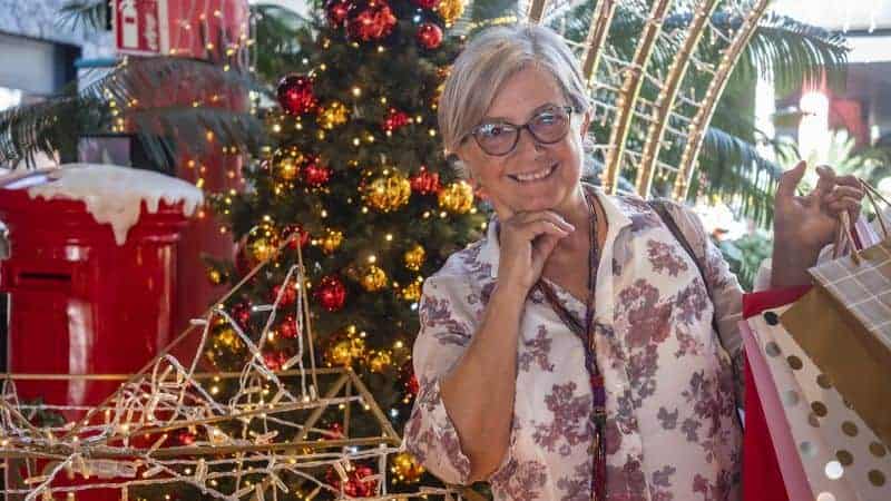 Χαμογελαστή πελάτισσα σε εορταστικό περιβάλλον κάνει τα χριστουγεννιάτικα ψώνια της, μπροστά από στολισμένο δέντρο και φωτεινές διακοσμήσεις.