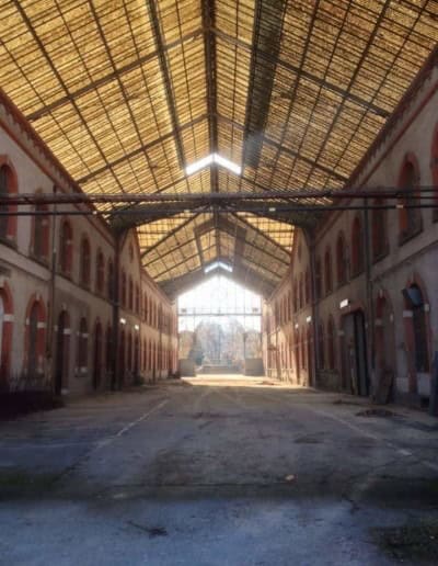 Interior view of an abandoned industrial building with a high, arched roof and natural light streaming through.