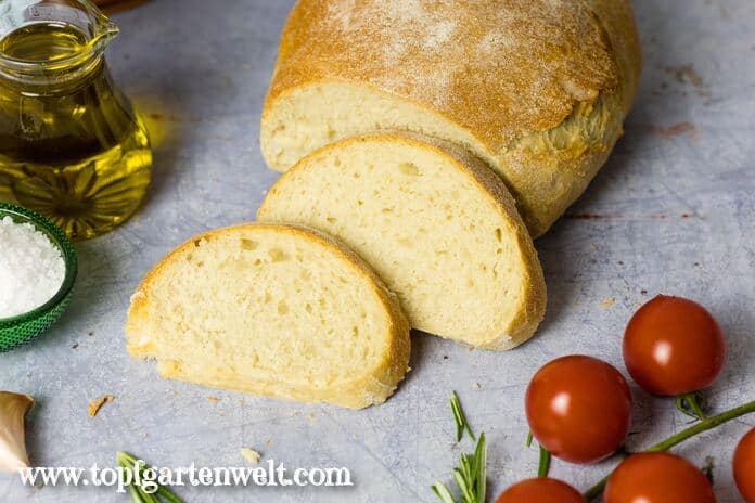 Ein Ciabatta ist angeschnitten. Zwei Scheiben liegen vor dem Brot. Im Hintergrund sind Olivenöl und Tomaten zu sehen.