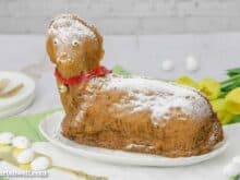 Ein Osterlamm aus Sandmasse steht auf einem weißen, ovalen Teller. Im Hintergrund sind gelbe Tulpen zu sehen.