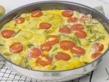 Frittata mit grünem Spargel und Kartoffeln in einer Pfanne, die auf einem schwarzen Abkühlgitter steht. Davor ist ein wenig frischer Rosmarin zu sehen.