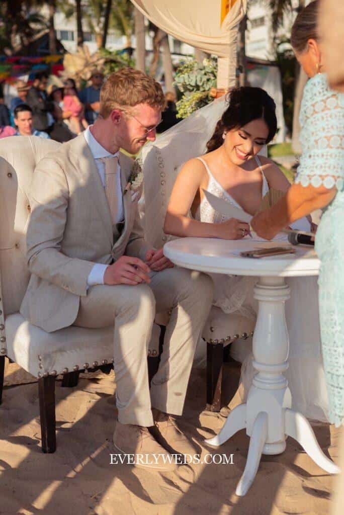 Bride and groom signing wedding documents on beach.