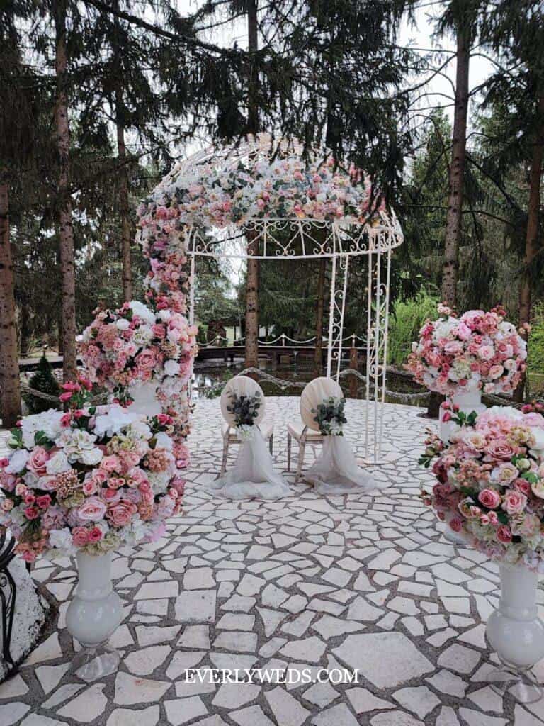 Pink and white floral arch with gazebo for budget outdoor wedding ceremony on stone patio with forest backdrop