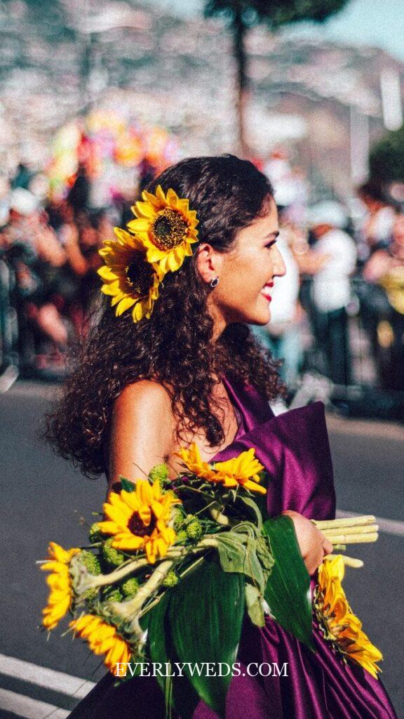 A woman in a purple dress holds a bouquet of sunflowers and has a large sunflower in her curly hair. She is smiling and standing outdoors at a lively event, with blurred people and decorations in the background.