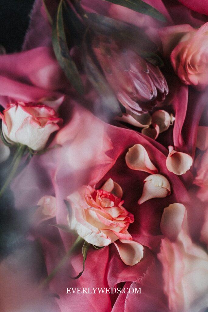 A close-up of pink and cream roses and calla lilies arranged on a deep pink fabric, with soft focus