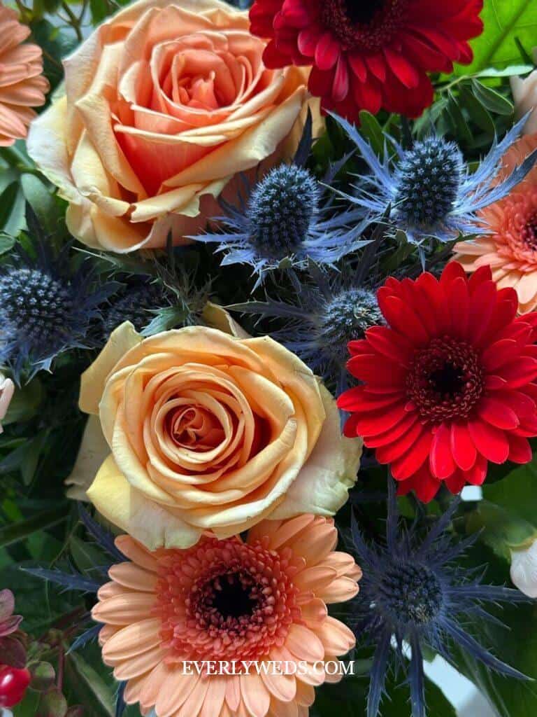 A close-up of a vibrant flower arrangement featuring peach roses, red and peach gerbera daisies, and spiky blue eryngium flowers, with green leaves in the background.