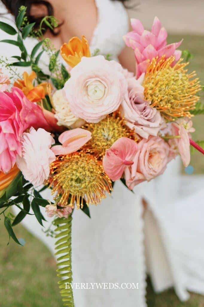 A bride holds a colorful bouquet with pink, orange, and yellow flowers, including roses and proteas, against a blurred outdoor background. 