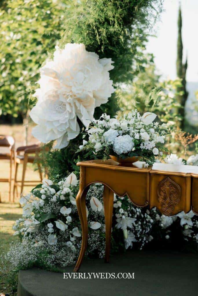 Outdoor wedding setup featuring a wooden table with floral carvings, surrounded by lush greenery. Large white paper flowers and fresh white floral arrangements decorate the scene.
