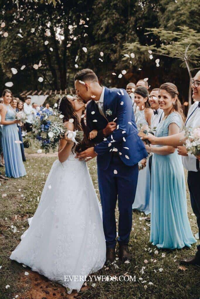 Couple kissing at outdoor wedding ceremony with flower petals falling, surrounded by bridesmaids in blue dresses