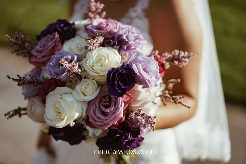 A bride holds a bouquet with white, lavender, purple, and burgundy roses, accented with pink sprigs. The background is softly blurred and the bride’s white lace dress is partially visible. 