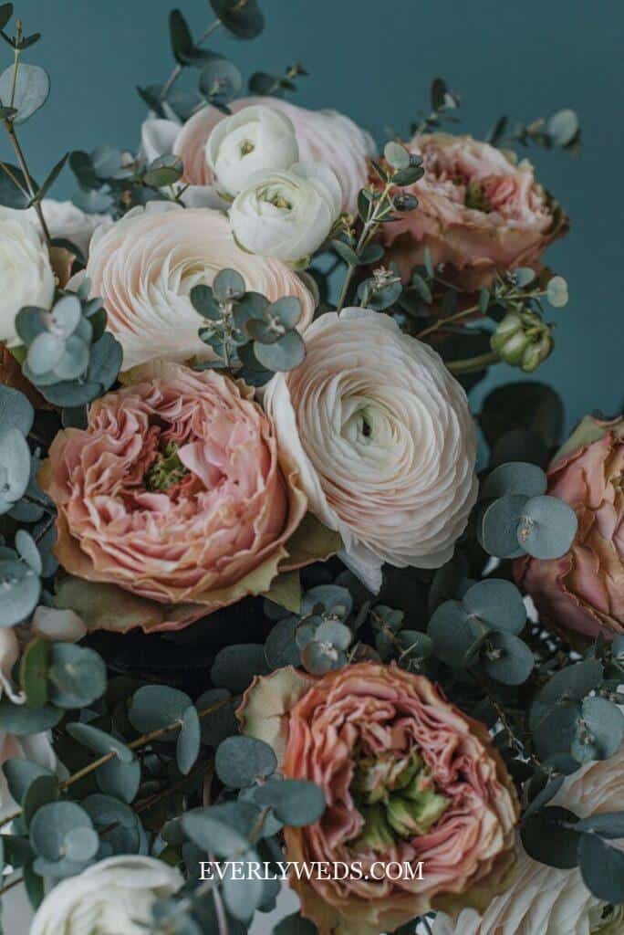A close-up of a bouquet featuring blush pink and white ranunculus flowers with eucalyptus leaves, arranged against a muted teal background. The petals are soft and layered.