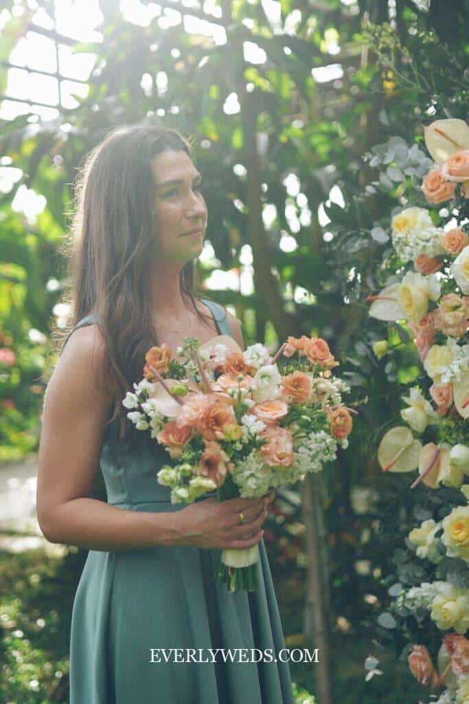 A woman in a sleeveless teal dress holds a bouquet of peach and white flowers, standing outdoors near a floral arrangement, with sunlight filtering through greenery.