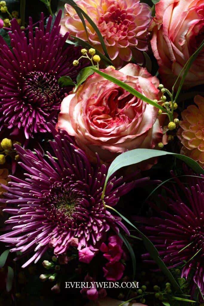 A floral arrangement with deep purple chrysanthemums, pink roses, peach dahlias, and green foliage, shown in soft lighting.