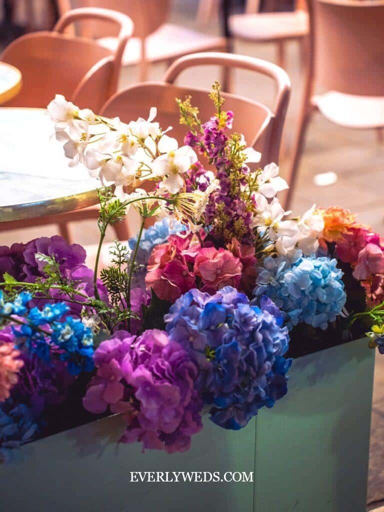 A decorative floral arrangement with vibrant blue, purple, pink, and white flowers sits in front of a table and chairs at an indoor venue.