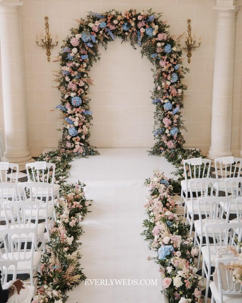 A floral wedding arch decorated with pink, blue, and white flowers stands at the end of an aisle lined with white chairs and matching flower arrangements. The elegant indoor setting has soft lighting and cream-colored walls.