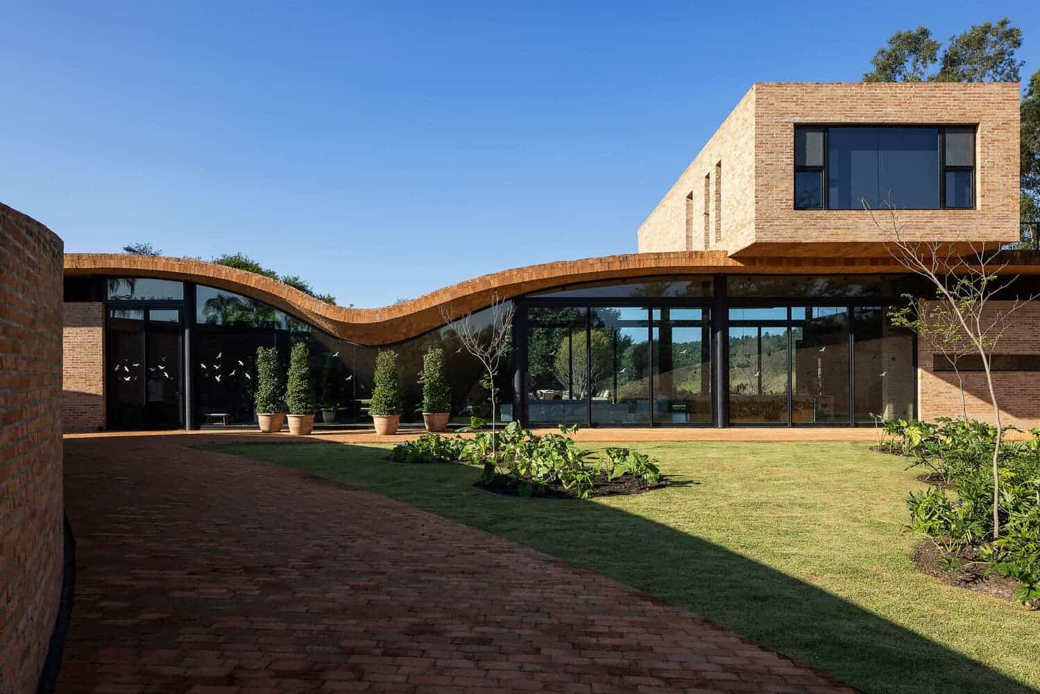Contemporary modern house with curved brick roof and large glass windows, surrounded by a landscaped garden and paved walkway, showcasing innovative architecture and design.