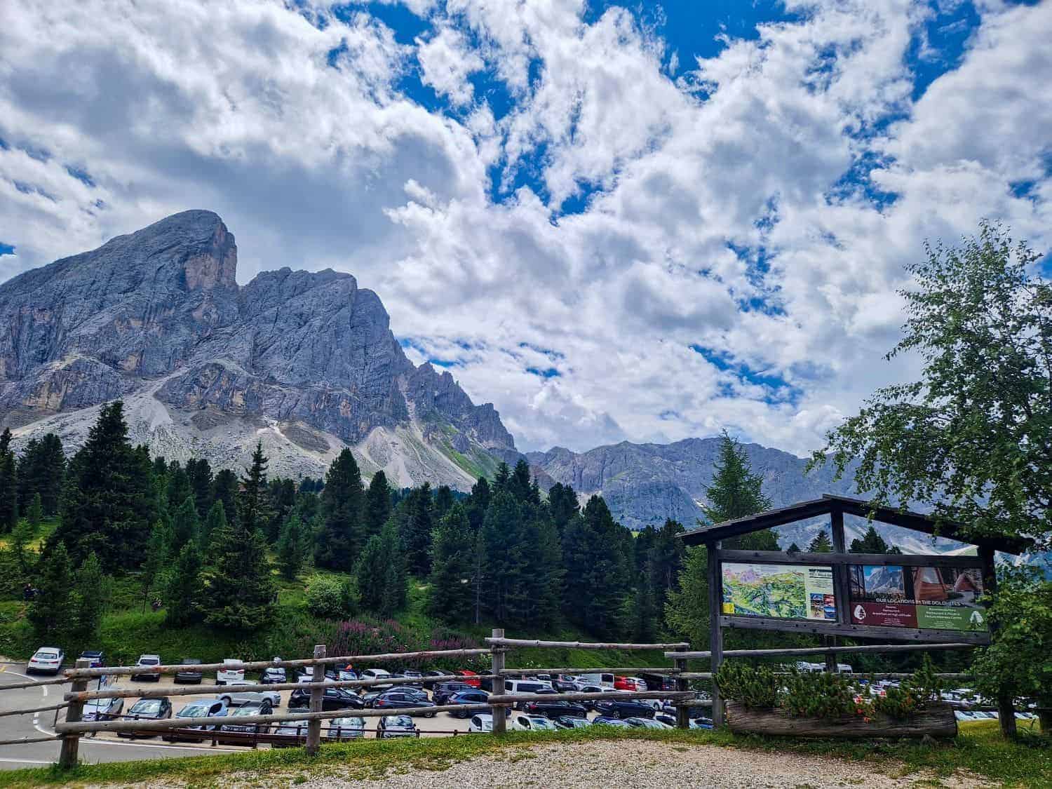 Scenic mountain view with rocky peaks and scattered clouds above. A parking area with numerous cars is surrounded by lush green trees. A wooden information board is in the foreground on the right.
