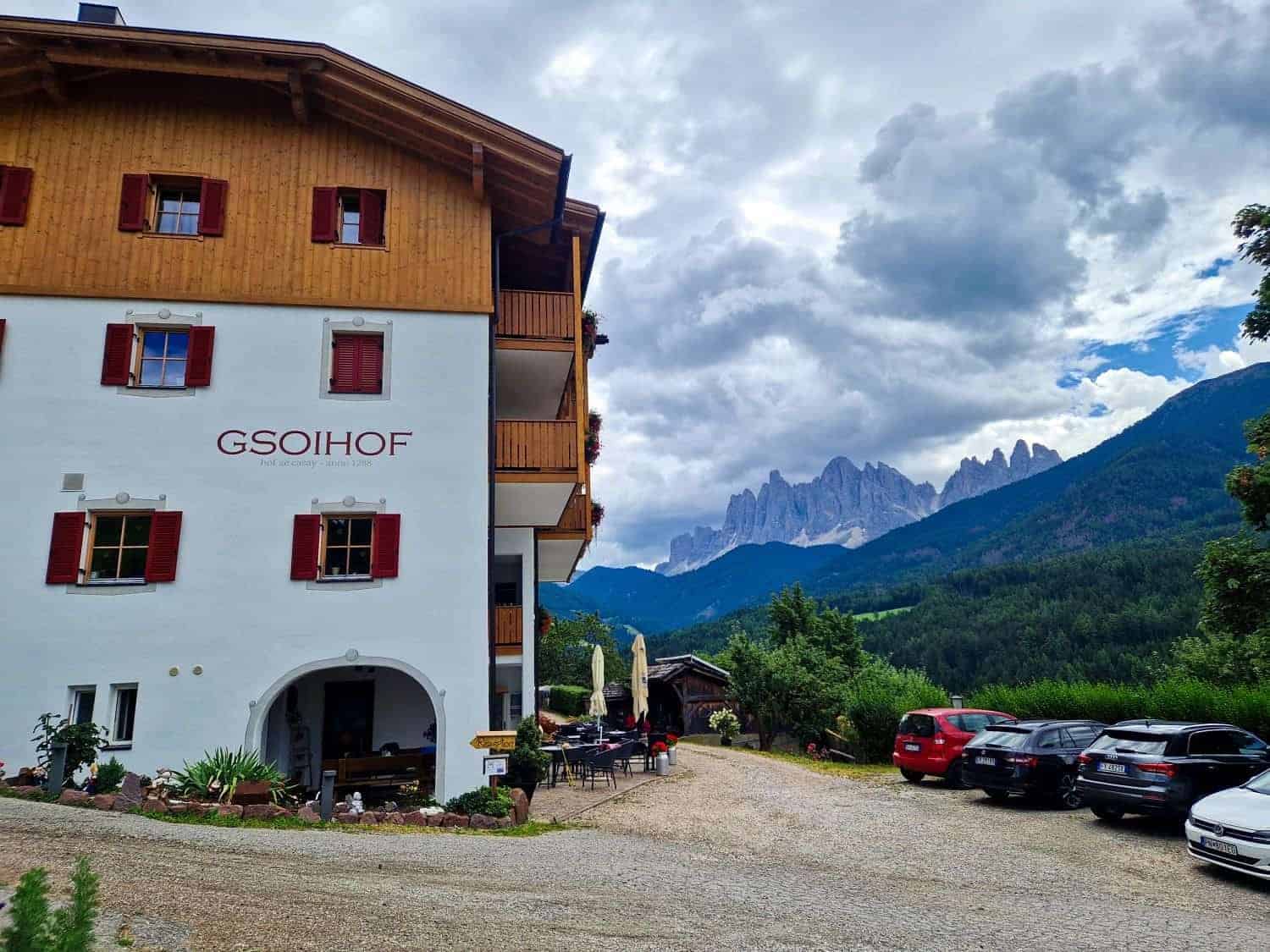 A rustic house labeled "Gsoihof" with red shutters stands against a mountainous backdrop. Cars are parked nearby on a gravel path, while dramatic clouds loom over distant peaks. A few outdoor tables and trees complete the tranquil scene.