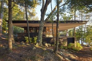Modern minimalist house with large overhanging roof in a forest setting, blending contemporary architecture with nature, featuring extensive glass walls and outdoor balcony.