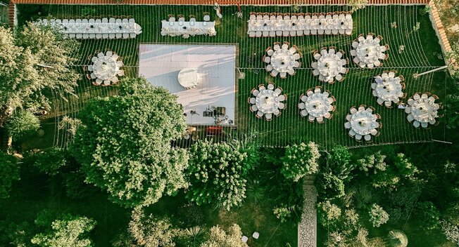 A long dining table with wooden chairs is elegantly set with glasses, plates, and flowers, overlooking a scenic landscape with mountains and trees in the background.